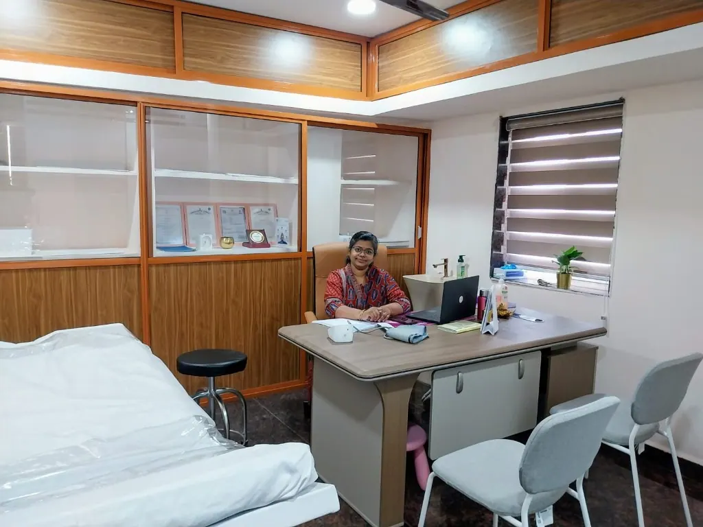 Dr. Krishna P Syam at her desk in the Cura Femina consultation room, with patient chairs, an exam table, and certificates in the cabinets behind her.