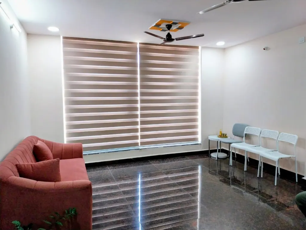 Waiting lounge at Cura Femina with a terracotta sofa, grey and white chairs, zebra blinds on a large window, ceiling fan, and reflective dark floor.